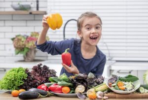 ילדה עומדת במטבח כאשר לידה מגוון של ירקות טריים והיא מחזיקה גמבה ופלפל בידיים